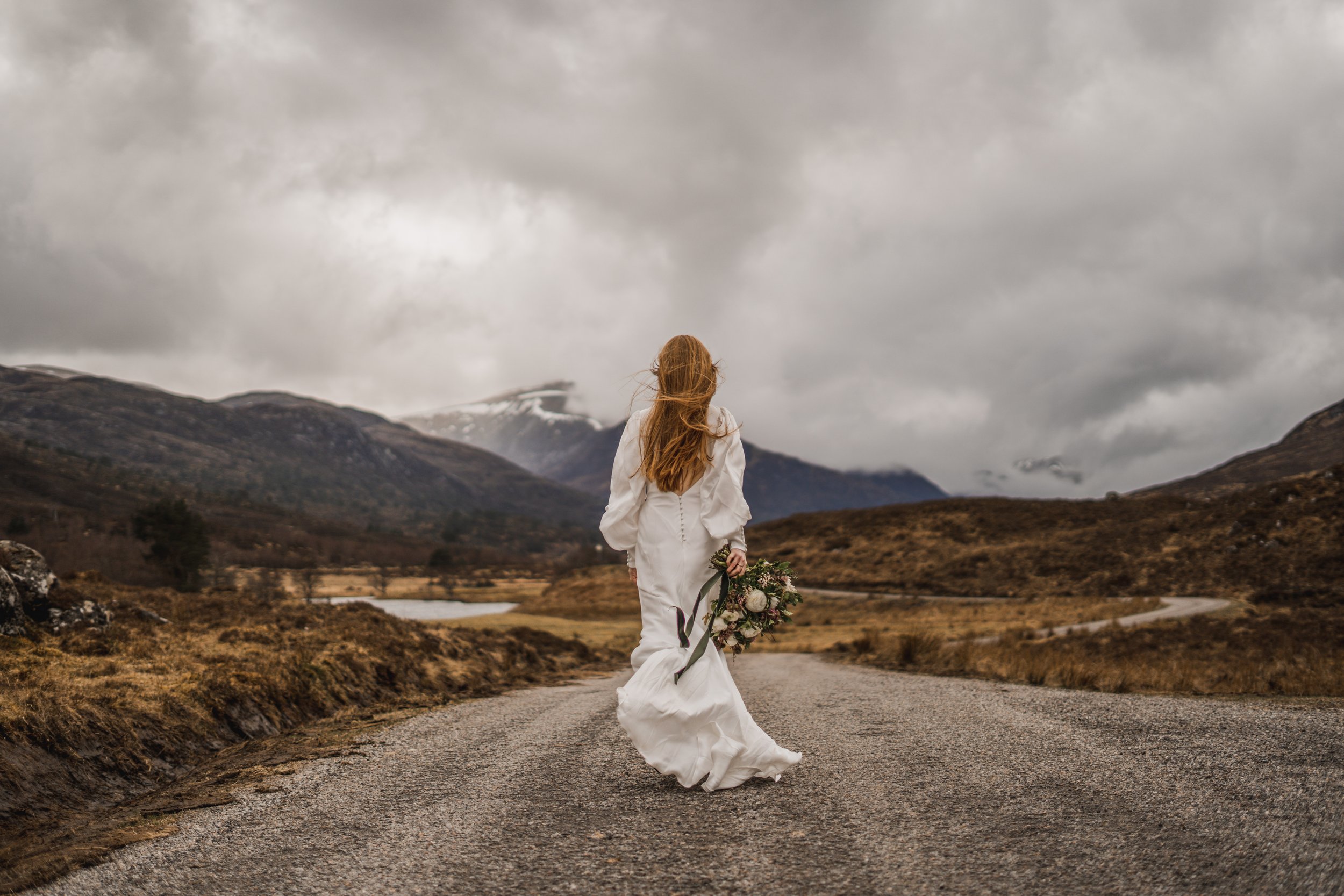 Bride walking along a country road with a bouquet in hand, surrounded by the Scottish hills